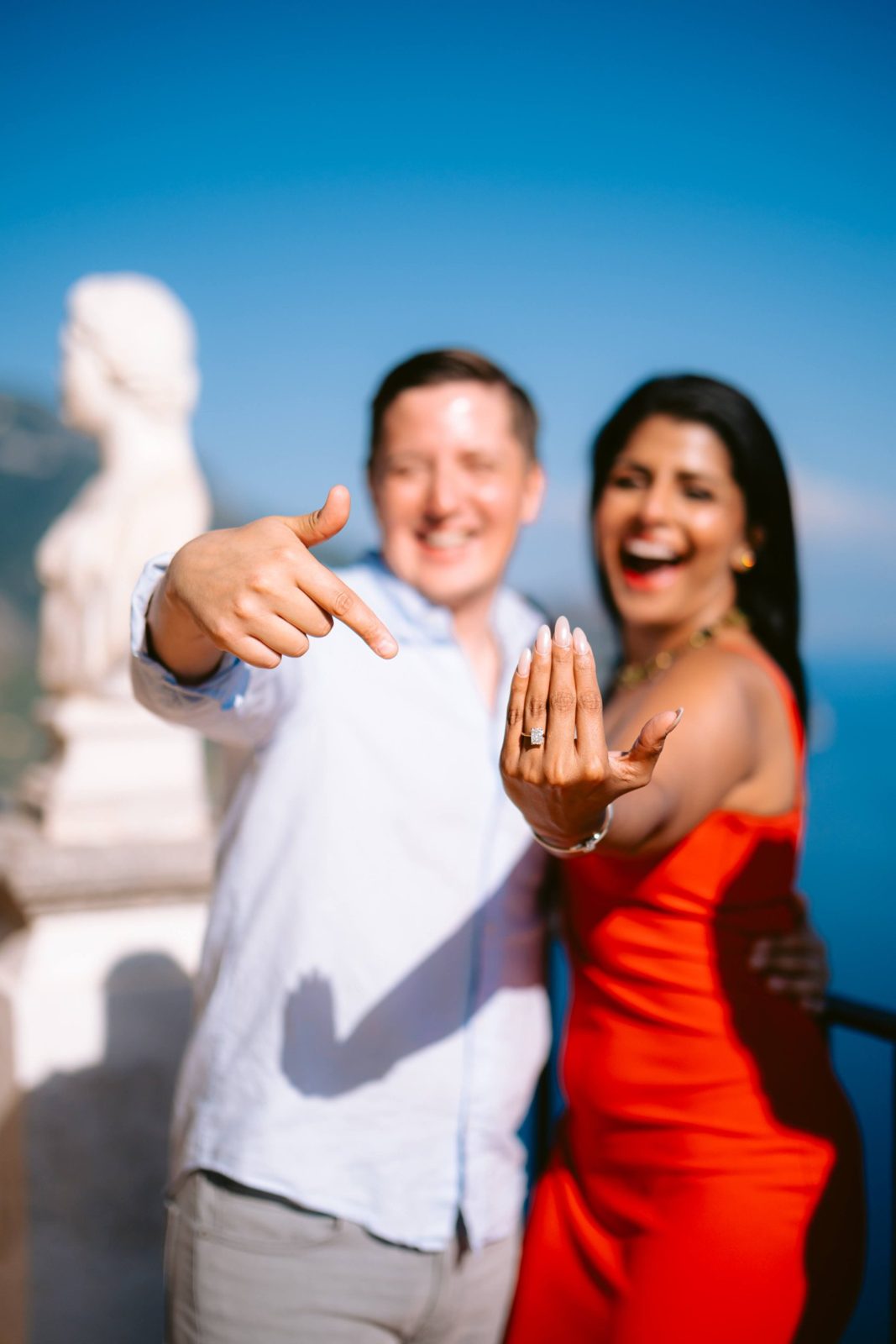 A laughing couple by the sea as she shows her engagement ring